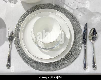 Place setting with shimmering placemat, white plates, bowl, knife, fork, and spoon on a tablecloth Stock Photo