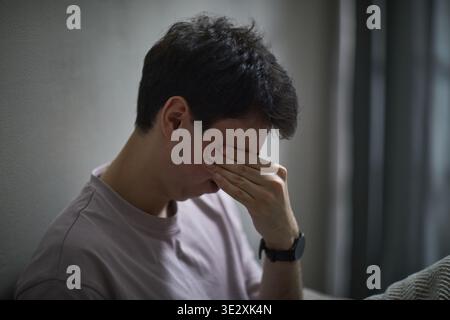 Young adult Caucasian man sitting against wall covering face with hand showing stressed expression, short dark hair visible, wearing wristwatch, conveying emotion Stock Photo