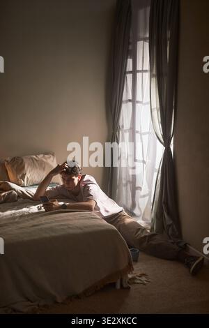 Vertical of Young adult Caucasian man sitting on floor beside bed holding smartphone, leaning against mattress with thoughtful expression, sunlight streaming through window, conveying emotion Stock Photo