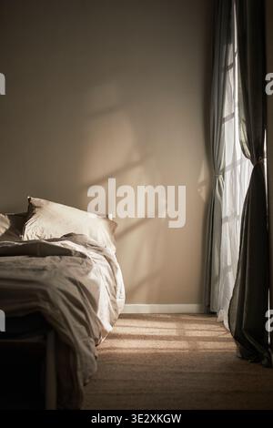 Vertical of Empty bedroom interior showing unmade bed with sunlight streaming through window, casting shadows on wall and floor, evoking sense of solitude and quiet emotional atmosphere Stock Photo