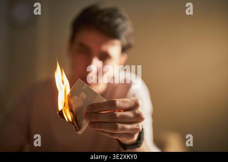 Young adult Caucasian man holding burning photograph of beloved woman, focusing on flames consuming image, expressing intense emotion and letting go of past memories in personal setting Stock Photo
