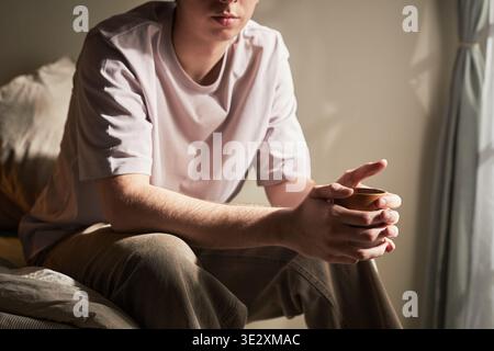 Young Caucasian man sitting on bed holding cup with both hands, looking down with thoughtful expression, sunlight streaming through window, conveying introspective emotion Stock Photo