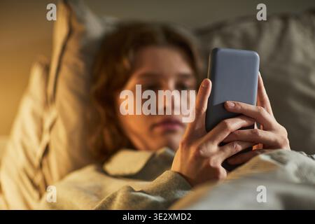 Caucasian young adult woman lying in bed holding smartphone with both hands, focusing on device screen, displaying worried facial expression, engaging with technology in private setting Stock Photo