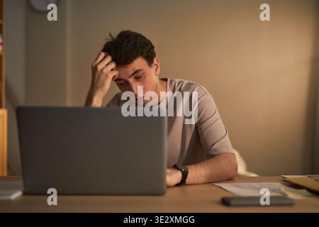 Young adult Caucasian man sitting at desk holding head in hand, while looking at laptop screen, showing stressed or worried facial expression in home office setting Stock Photo
