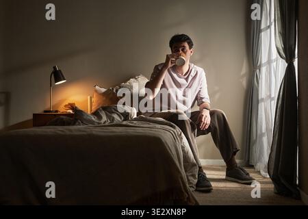 Young adult Caucasian man sitting on bed drinking from mug, gazing forward with neutral expression, morning light streaming through window, conveying emotion of contemplation or introspection Stock Photo