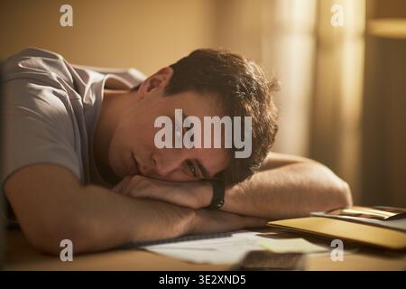 Portrait of Caucasian young adult man resting head on arms at desk, gazing into camera with tired expression, surrounded by paperwork and smartphone, conveying emotional fatigue Stock Photo