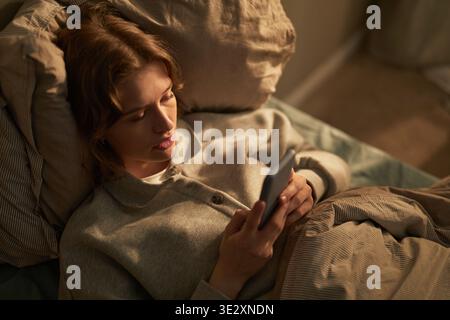 Caucasian young adult woman lying in bed using smartphone, displaying thoughtful facial expression while reading or texting, head resting on pillow, focused on device Stock Photo