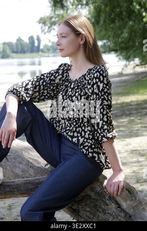 Fashionable Woman Relaxing by River in Flowy Black and White Blouse and Denim Trousers – Casual Lifestyle and Natural Summer Fashion Concept Stock Photo