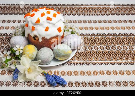 A beautifully decorated Easter kulich sits on a plate surrounded by colorful, hand-painted eggs and delicate spring blossoms, all set against a richly Stock Photo