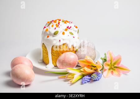 A festive Easter kulich cake adorned with white icing, dried fruits, and seeds, surrounded by pastel-colored eggs and blooming tulips and muscari flow Stock Photo
