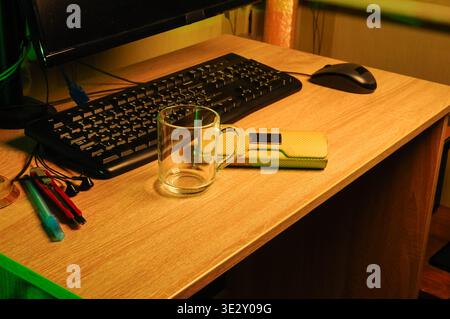 A close up view of a wooden desk surface featuring a keyboard, mouse, empty glass mug, earbuds, and writing tools Stock Photo