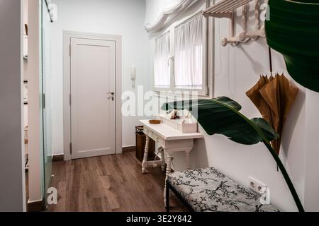 Modern home entryway with white wall, wooden floor, a decorative table, bench, coat rack, and a large plant Stock Photo