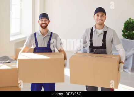 Portrait of two smiling movers in uniform holding cardboard boxes, assisting in relocation process. Stock Photo
