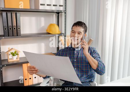 A young architect is standing at his desk while discussing a project on the phone, carefully reviewing construction plans with focus and attention in Stock Photo