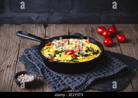 Frittata with spinach and tomato in a cast iron pan on wooden background, copy space Stock Photo