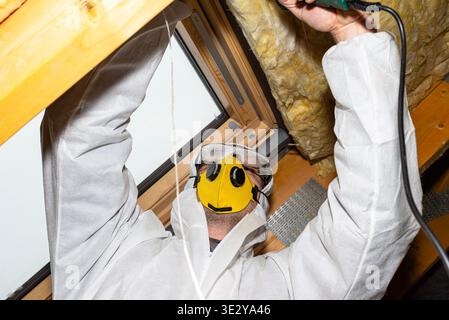 Man in protective clothing and mask working on attic insulation and roof window Stock Photo
