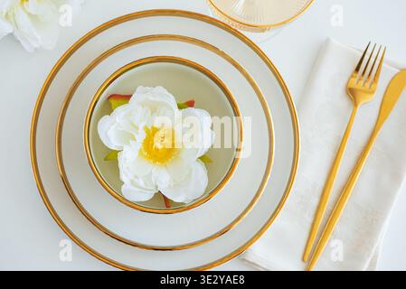 Elegant universal table setting featuring stacked white ceramic dishes with gold accents, a neatly folded neutral napkin, and gold cutlery arranged in Stock Photo