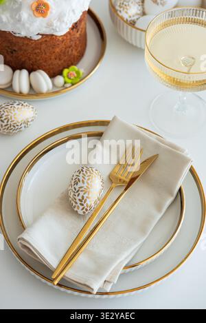 Elegant Easter table setting featuring white plates with gold trim, a folded napkin, gold cutlery, Easter cake and decorative egg Stock Photo