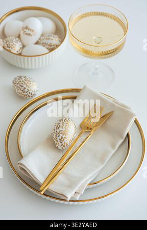 Elegant Easter table setting featuring white plates with gold trim, a folded napkin, gold cutlery, and decorative egg on white table Stock Photo
