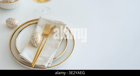 Elegant Easter table setting featuring white plates with gold trim, a folded napkin, gold cutlery, and decorative egg on white table Stock Photo