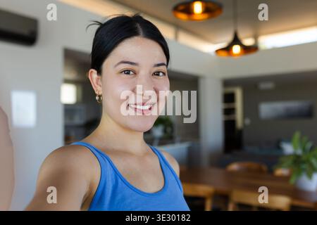 Asian woman taking selfie, smiling in home kitchen in blue tank top with smartphone, potted plant Stock Photo