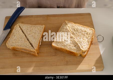 Wooden board holding two wheat sandwiches and blue knife on white counter, light casting shadow Stock Photo