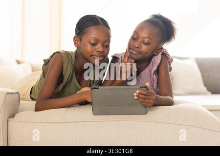 African American sisters, youth, child lying on sofa in living room using tablet in folio case Stock Photo