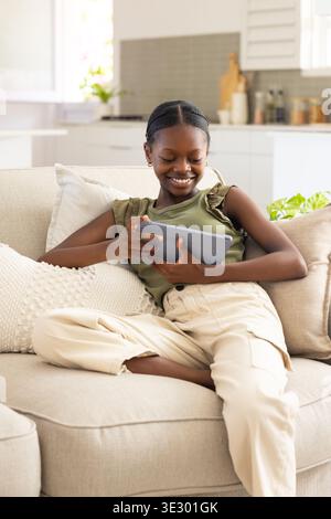 Gray tablet resting on light beige sofa and showing content near window with kitchen background Stock Photo