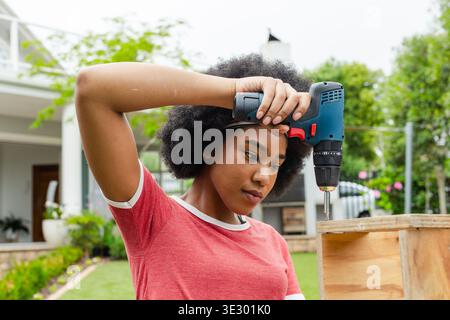 African American woman wearing red tee dark headband using drill on wooden crate in backyard Stock Photo