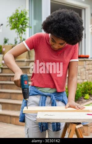 African American woman drilling wooden plank on portable workbench using cordless drill on porch Stock Photo