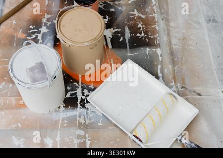 Displaying paint cans and tray sitting on protective plastic sheet at garage, with roller and stick Stock Photo