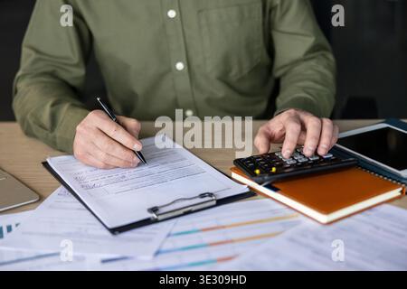 Person working on a desk, calculating expenses with a calculator and writing on official documents, managing personal or business finances, taxes, and accounting Stock Photo