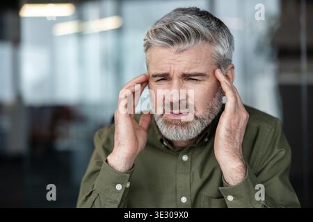 Distressed mature man feeling intense headache and migraine pain, touching temples with both hands while experiencing pressure and stress from work or discomfort Stock Photo