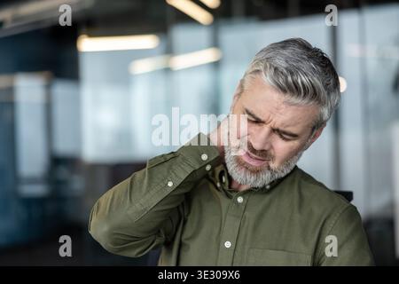Mature businessman with gray hair and beard rubbing his neck in a modern office, grimacing from cervical pain and poor posture while working at a computer, stressed and fatigued Stock Photo