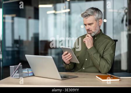 Experienced businessman with grey hair studying data on a digital tablet at his desk, laptop open, focused on strategy, problem solving and planning for a corporate project Stock Photo
