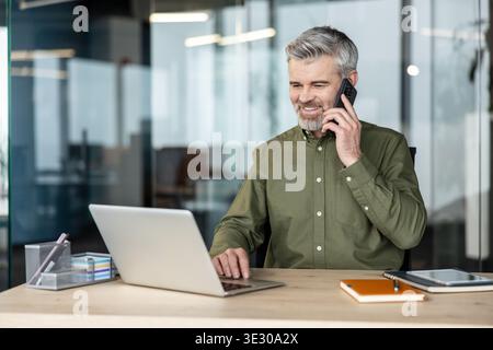 Happy mature man with a beard and gray hair working on a laptop at his desk in a modern office, communicating on a smartphone with a client or colleague Stock Photo