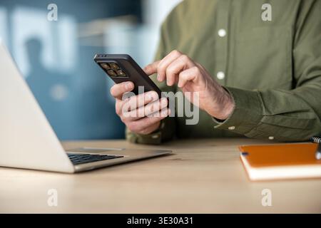 Man using smartphone with both hands, scrolling at an office desk beside laptop and notebook, focused on browsing and digital communication in a modern workspace setting Stock Photo