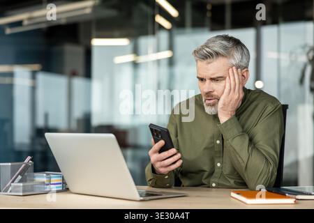 Mature man sitting at an office desk, looking distressed while checking his mobile phone, experiencing stress or anxiety from a difficult message or result Stock Photo