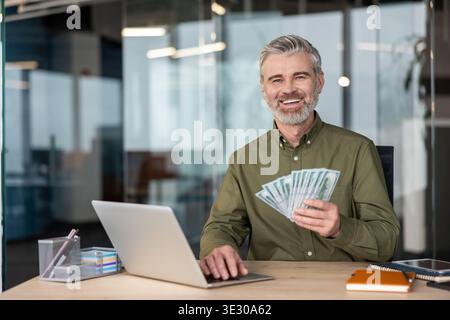 Happy mature businessman holding cash money and using a laptop, celebrating successful financial profits and online earnings while working from an office Stock Photo