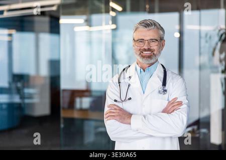 Smiling confident middle-aged male doctor with a grey beard and glasses wearing a white lab coat and stethoscope, posing with arms crossed in a modern doctor's office Stock Photo