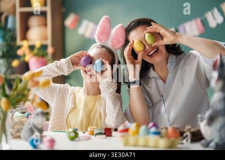 Joyful mother and daughter enjoying Easter festivities, wearing bunny ears and holding decorated eggs playfully in front of their faces. The bright an Stock Photo
