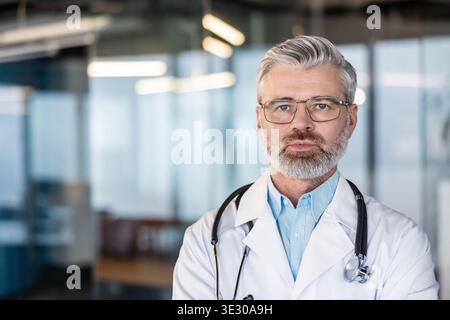 Experienced male doctor wearing a white lab coat, blue shirt, and stethoscope standing confidently in a modern clinic or hospital setting, representing professional healthcare expertise Stock Photo