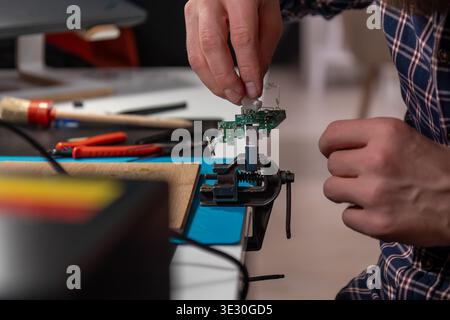 A skilled technician meticulously works on a small electronic circuit board clamped in a metal vise, using a hot air rework station and tweezers to pe Stock Photo