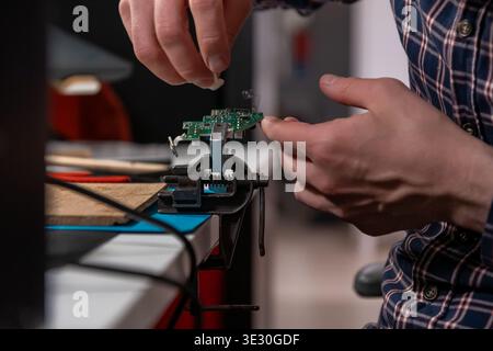 A skilled technician meticulously works on a small electronic circuit board clamped in a metal vise, using a hot air rework station and tweezers to pe Stock Photo