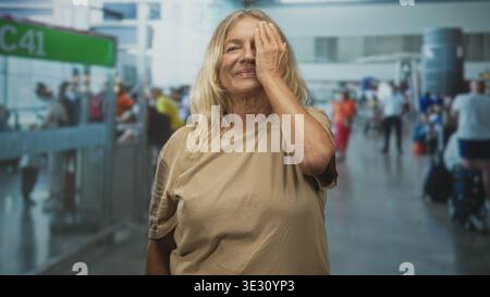 Senior woman with blonde hair covers one eye with her hand while smiling in a tan tee at busy airport gate c41 among travelers and luggage; serenity. Stock Photo