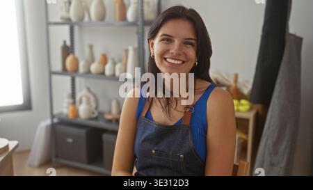 Woman adjusts hair with a smile while wearing apron in studio surrounded by pottery vases on shelves; creative joy. Stock Photo