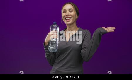 Woman holding a large plastic water bottle and waving right hand in studio with purple backdrop, smiling at camera; hydration joy. Stock Photo