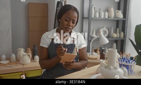 Woman potter in apron writes in a notebook with hands holding pencil beside ceramic vases on a studio workbench; concentration. Stock Photo