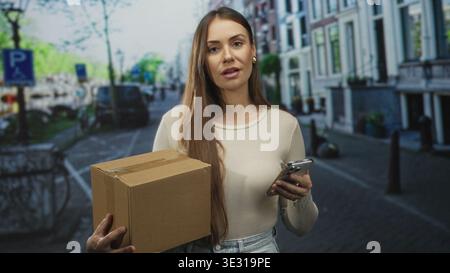 Woman holding cardboard box and smartphone looks at phone while standing on an urban street with parked cars and bicycles; focused delivery. Stock Photo