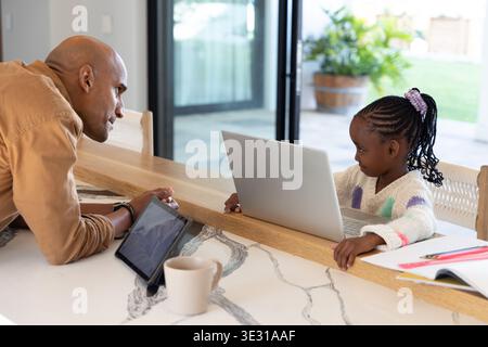 African American man and girl leaning over counter at breakfast bar using silver laptop, copy space Stock Photo